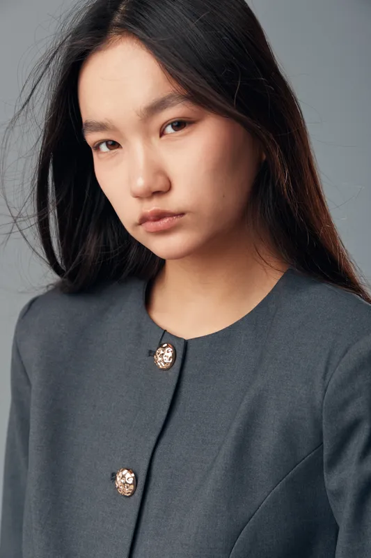 Young woman with long black hair and natural makeup wearing a gray buttoned blazer posing in studio with neutral expression and plain background for portrait concept.