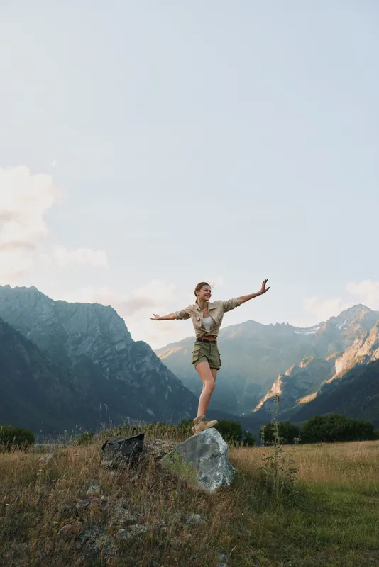 A young woman in a khaki shirt and shorts stands on a rock with arms outstretched enjoying the mountain landscape under a clear sky during daytime.