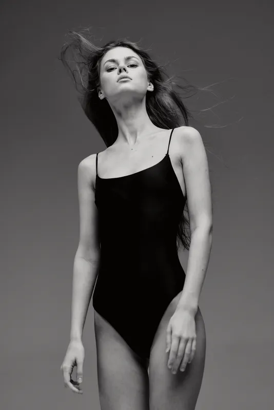 Black and white portrait of a tall woman in a black swimsuit with windblown hair and confident expression
