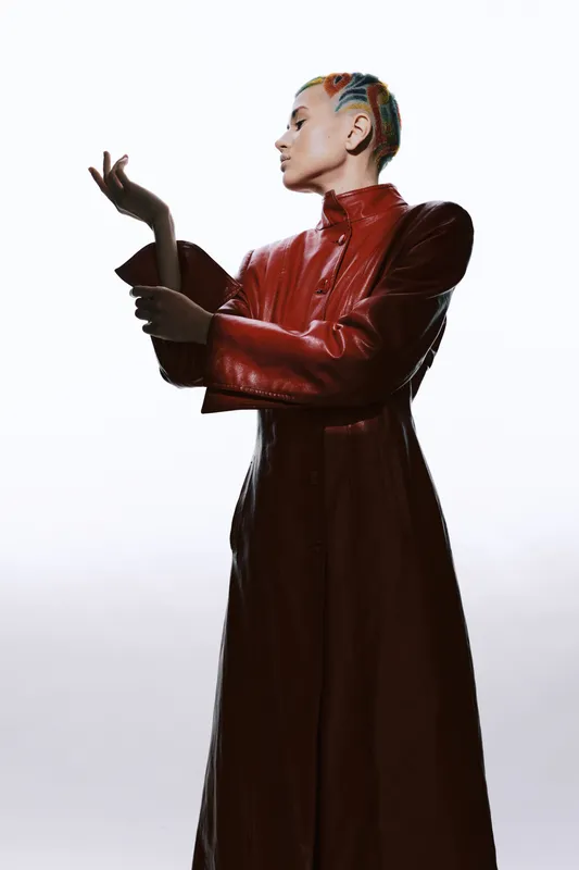 woman in red leather coat with colorful braided hair stands confidently in profile against white background, showcasing modern stylish fashion and bold attitude