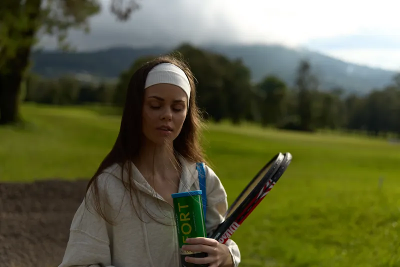Young female athlete in casual sportswear holding tennis rackets and a drink in a serene outdoor setting, embodying a sporty lifestyle and healthy habits