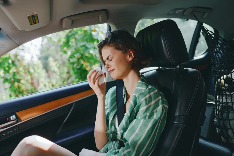A woman sits in a car seat wearing a seat belt, gazing to the side while touching her face, captured in soft natural light.