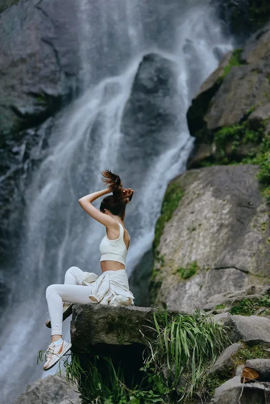 Woman sitting on rock by waterfall in sporty outfit, enjoying nature and peaceful outdoor environment with lush greenery and flowing water background.