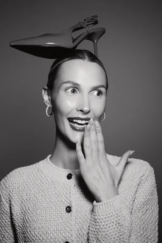 Caucasian woman with fair skin and dark hair holds hand near mouth with surprised smile, balancing high heel shoe on head. Concept of fashion and humor.
