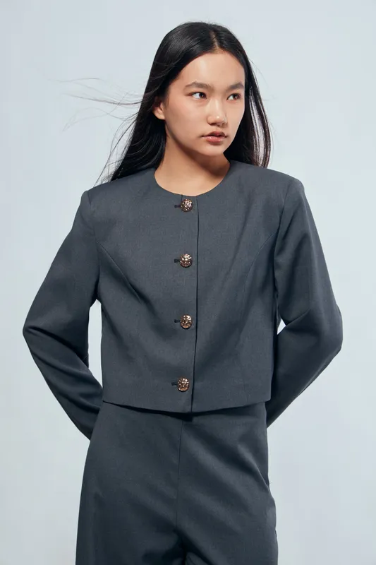 Young woman with long dark hair wearing gray formal suit with decorative buttons standing against plain background. Female model with confident pose and minimal makeup for business style.