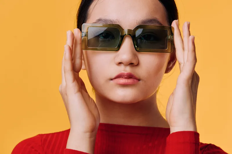 Young woman with dark hair wearing large square sunglasses and red turtleneck sweater standing against orange background looking at camera with neutral expression and hands near face.