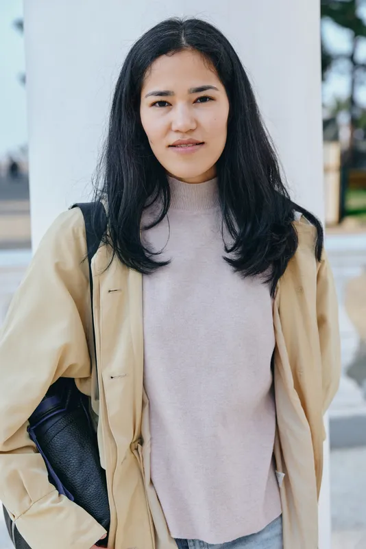 Woman in casual jacket and sweater stands outdoors with a focused expression, showcasing street fashion and confident style in an urban setting.