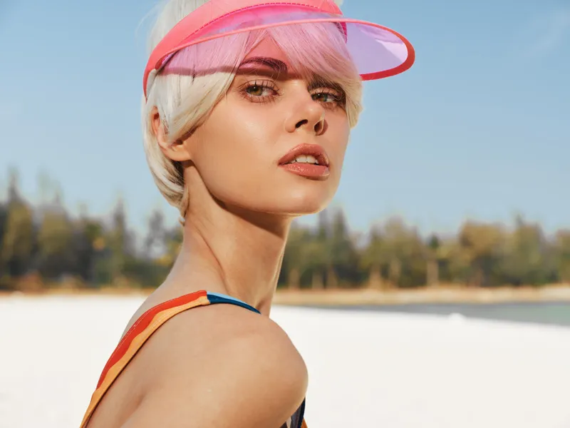 Portrait of a model on a sunny beach wearing a pink visor and swimsuit; warm sunlight highlights features, blue sky, soft background, carefree summer mood, vibrant colors.