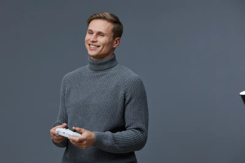 Smiling young man holding game controller in hands, wearing cozy gray turtleneck sweater, looking happy, isolated against plain gray background. Casual lifestyle concept