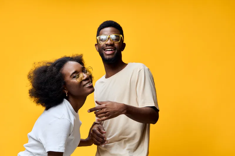 Joyful young couple laughing together on a vibrant yellow background, wearing casual summer clothing and stylish sunglasses, conveying happiness and warmth