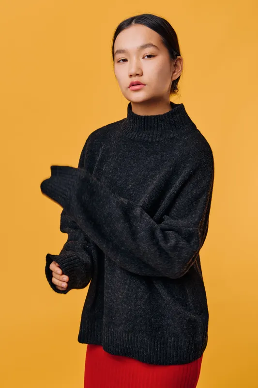 Young woman in black oversized sweater standing against orange background with serious expression, long hair tied back, and red skirt visible below. Fashion, portrait, minimal studio concept.