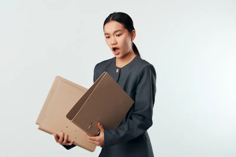Businesswoman with surprised expression holding cardboard folders in office for documents organization, paperwork, and professional work tasks on white background.