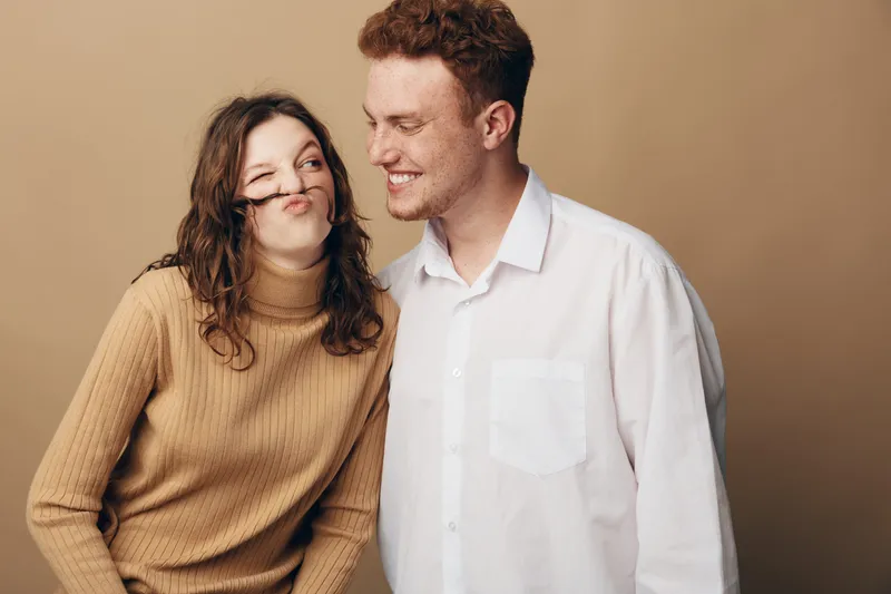 Playful young couple with curly hair smiling and making funny faces together against beige background. Concept of friendship, joy, and casual lifestyle in studio portrait.
