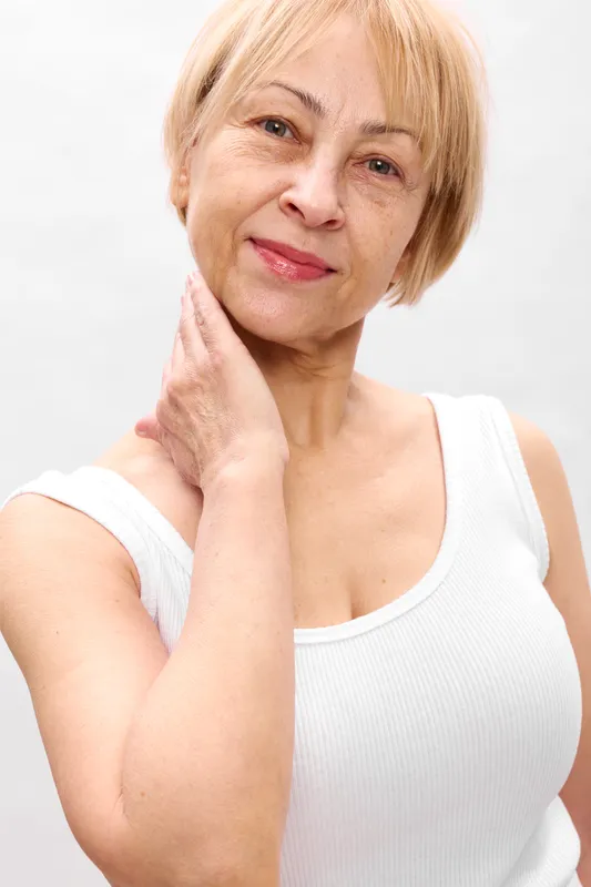 Elderly woman portrait in studio, blonde hair, gentle smile, hand on chin, senior age, white sleeveless top, calm expression, soft light, background, close up shot