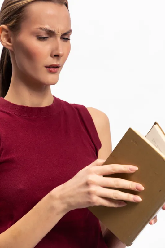 Confused woman in red sleeveless top reads old book with furrowed brow isolated on white background. Concept of puzzlement, curiosity, and vintage literature study.