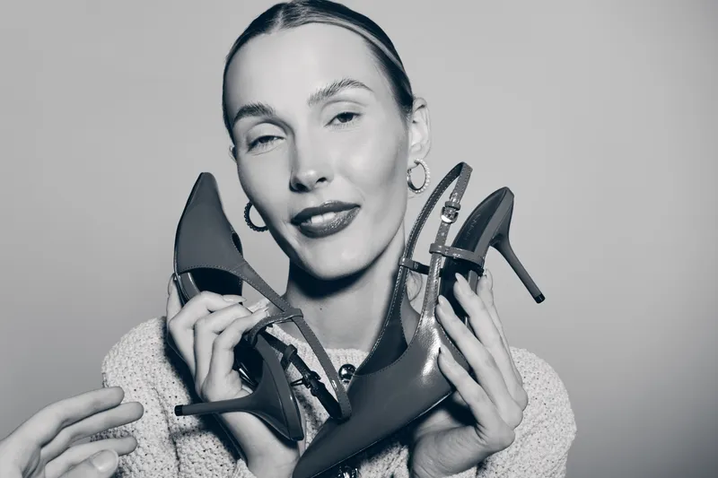 Young white woman with fair skin holds two black high heel shoes near her face with a confident smile in a studio portrait. Concept of fashion, style, and footwear.