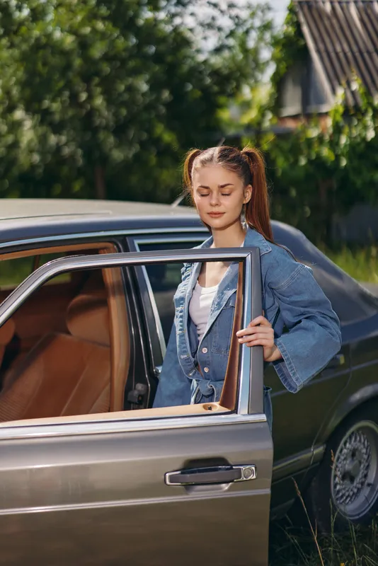 Young woman in denim jacket entering vintage car, showcasing casual style and a joyful spirit in natural outdoor setting.