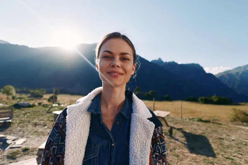 Woman portrait with smile in mountains at sunrise outdoors, nature traveler wearing denim and shearling jacket, happy young adult enjoying rural landscape and fresh morning light