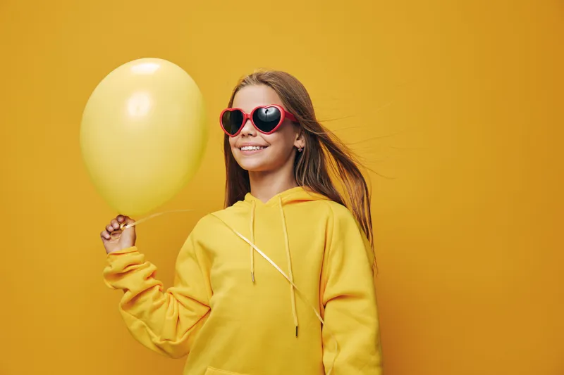 Girl in yellow hoodie and red heart-shaped sunglasses holding yellow balloon with smile in studio on yellow background. Child fashion, joy and party celebration concept.