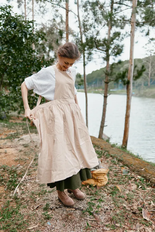 Young woman wearing a beige apron and green skirt, smiling by a riverside in nature, showcasing a relaxed, eco friendly lifestyle for spring or summer