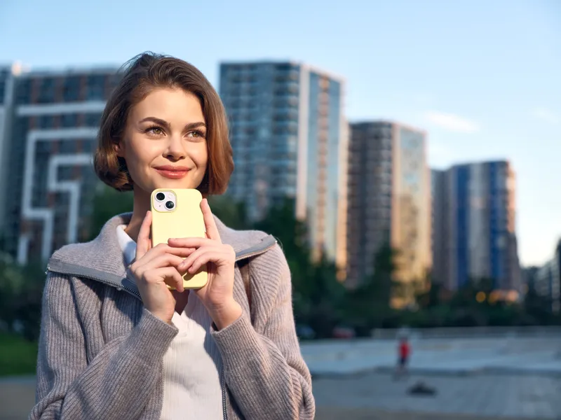 A cheerful woman stands outdoors in a modern city setting, smiling as she looks at a smartphone. The scene exudes pace, confidence, and a positive, productive business mood.