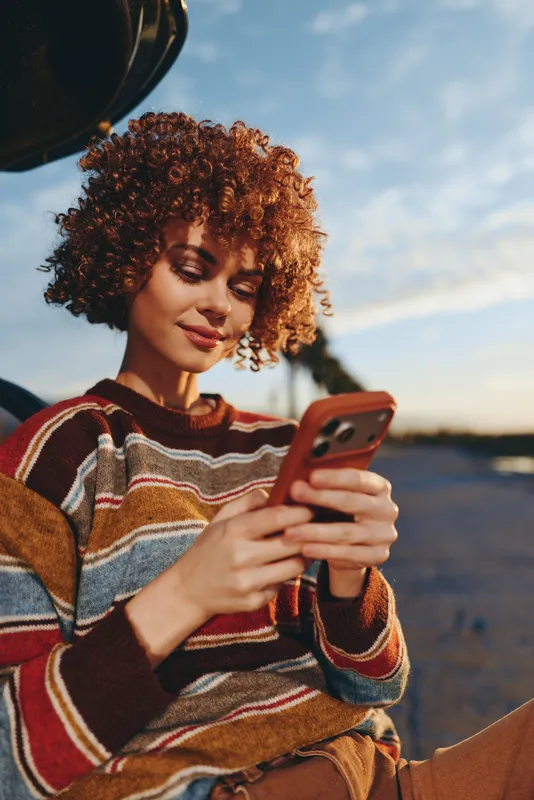 Woman lifestyle outdoors, curly hair woman with smile, holding smartphone, warm sunset evening light, striped sweater, relaxed casual pose, open sky background, soft breeze, calm