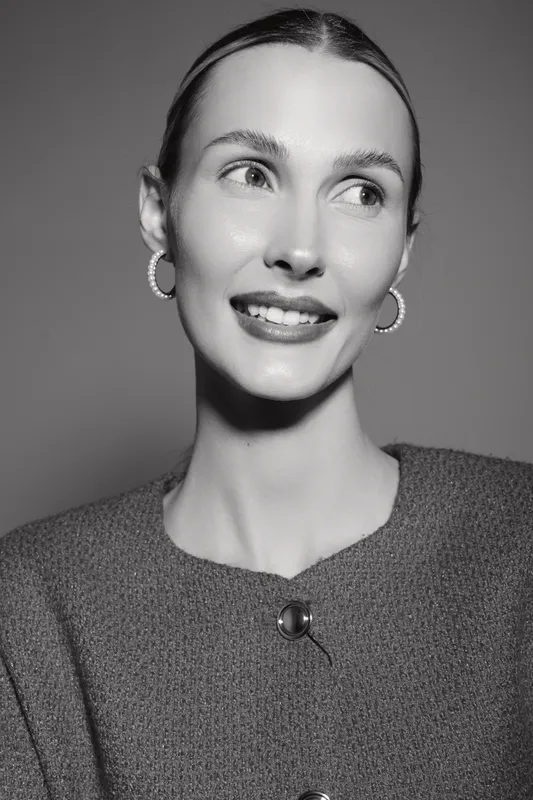 Elegant white woman with pale skin wearing textured sweater and hoop earrings looks aside smiling softly in monochrome portrait.