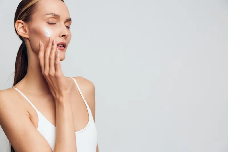 Young woman applies moisturizer to her face in a calm indoor studio, showcasing facial skincare and self care with soft light and a plain background, reflecting a natural beauty routine