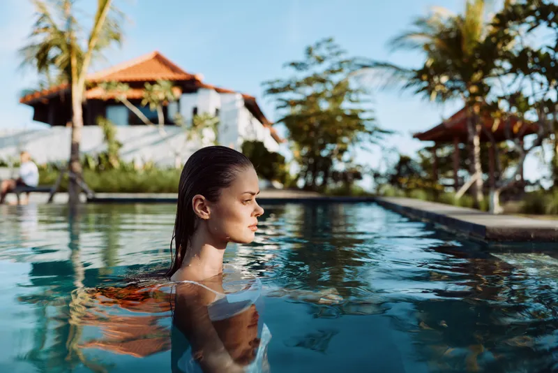 A woman floats in a clear pool, sunlight warming her skin as palm trees frame the resort area, conveying a calm lifestyle moment and vacation vibe in a bright outdoor setting.