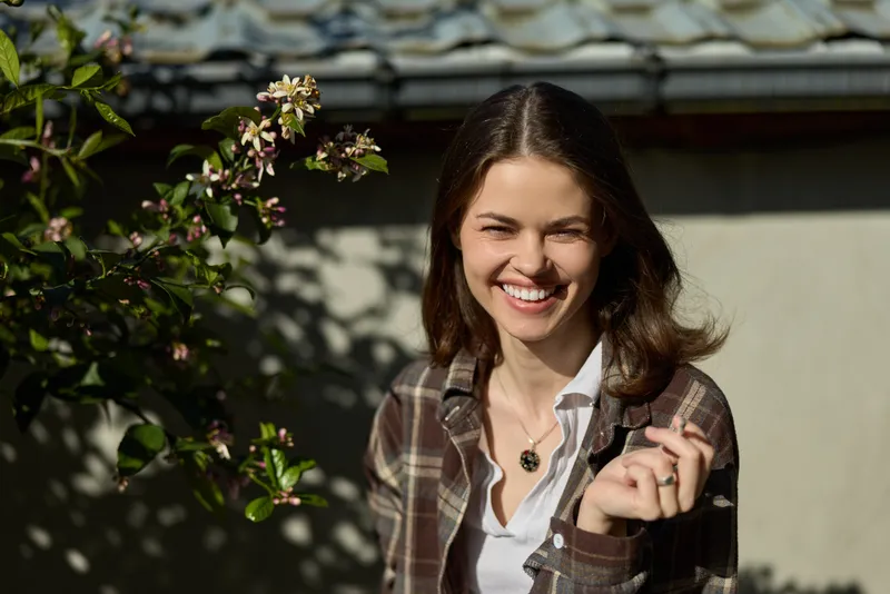 Smiling woman with long brown hair wearing a plaid shirt over a white top in an outdoor garden setting. Natural light creates a friendly, relaxed mood for lifestyle and candid moments