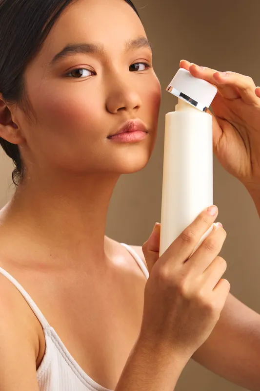 asian woman applies skincare product in a neutral studio, her calm expression highlighting a focused beauty routine and a serene moment of personal care.