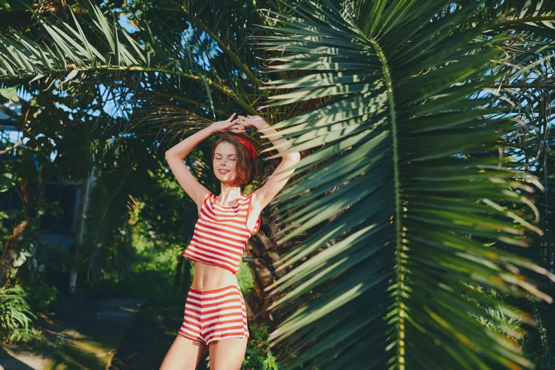A cheerful girl in a red and white striped outfit poses playfully among tropical palm leaves. Bright sunlight creates a carefree outdoor vibe, highlighting summer fashion and youthful energy.