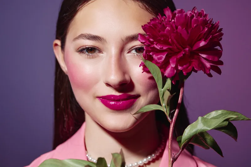 Young woman with bright makeup and shiny pink lipstick holding a burgundy flower close to her face against purple background. Concept for beauty, fashion, and creative portraits.
