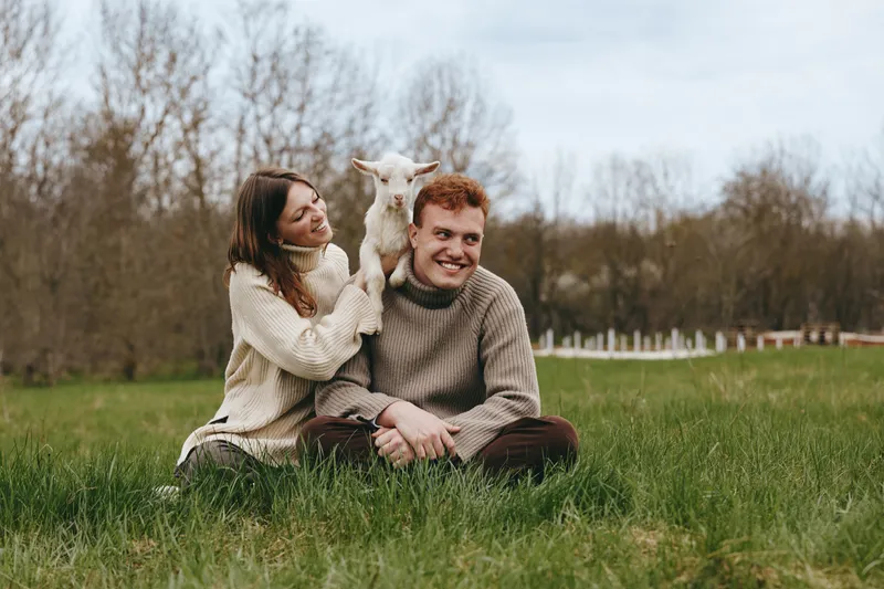 Smiling couple sitting on grass in a meadow with a playful goat kid resting on a shoulder. Cozy sweaters, natural light, rural lifestyle, simple happiness.