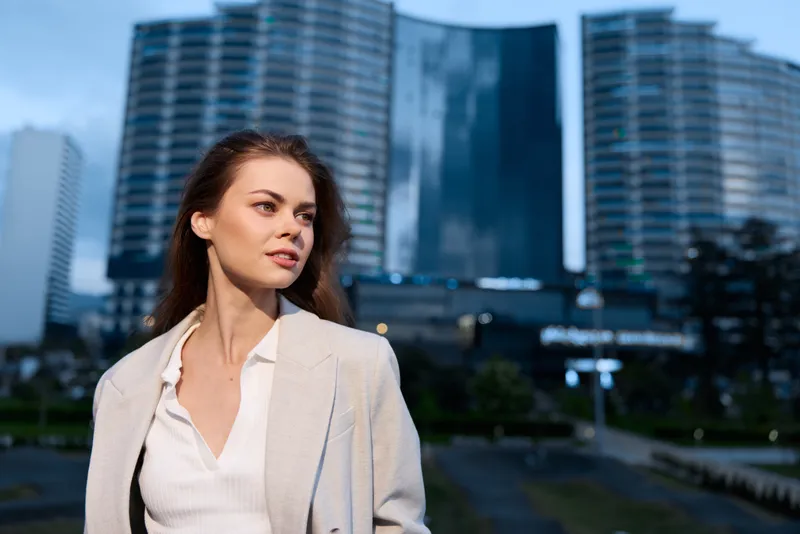 Confident woman standing outdoors in modern cityscape wearing light coat and white blouse with serious expression at dusk