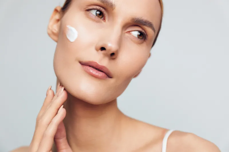 Close up of a young woman with light skin applying facial cream on her cheek during a skincare routine, looking calm and focused in a bright studio with soft lighting and a natural glow