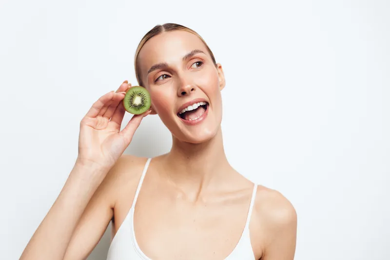 Young woman holding a fresh kiwi slice near her face smiling with open mouth, natural skin, white background, healthy lifestyle, bright lighting, casual wear, cheerful mood