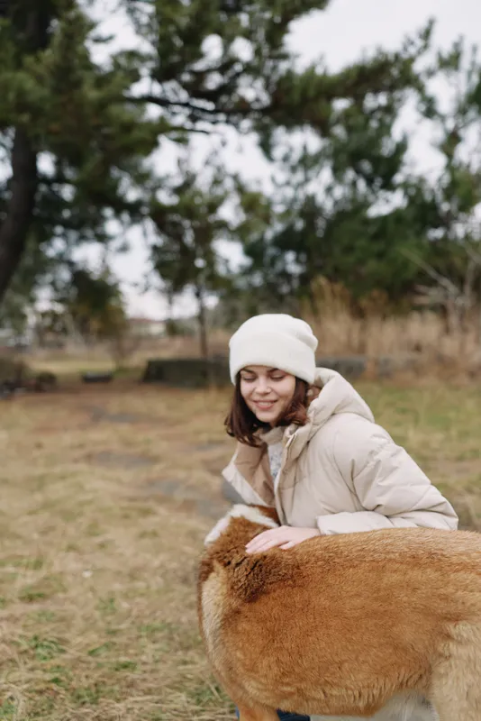 Girl in a warm beige coat and white beanie outdoors, smiling as she pats a fluffy alpaca in a grassy field on a cool day, sharing a wholesome nature moment