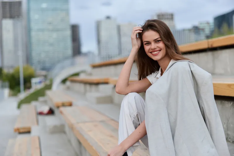 Smiling woman in light suit relaxing outdoors on city bleachers with modern buildings background and soft natural light, casual fashion style, positive emotion