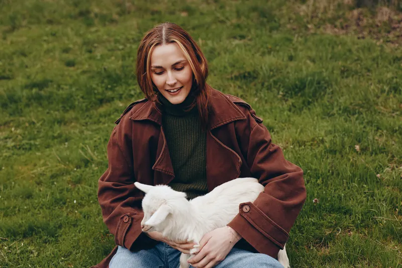 A woman in a brown coat sits on grass, gently embracing a goat kid. Outdoor lifestyle moment capturing calm bond, nature, and animal companionship in a rural setting.