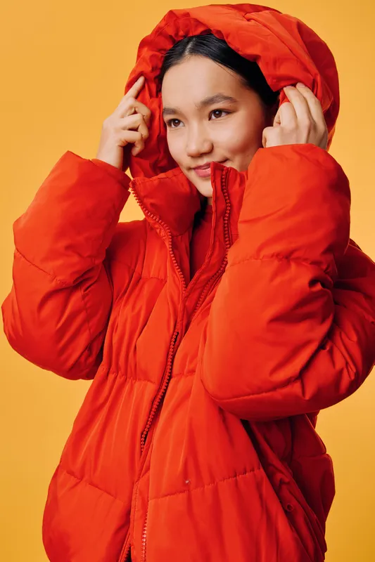 Woman in bright orange puffer jacket adjusting hood against yellow background. Portrait of young female model wearing warm winter coat with thoughtful expression and casual style.