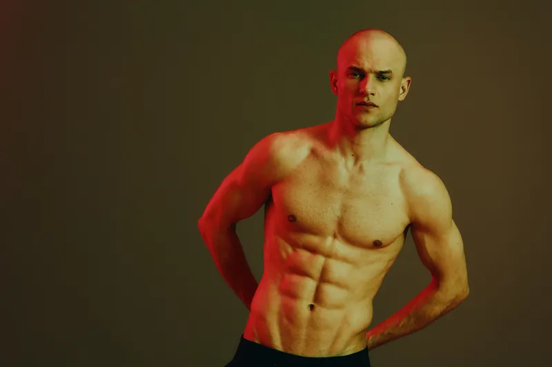 Fitness man with toned body posing in contrasting light, showcasing strength and determination, highlighting muscle definition and athleticism in a vibrant setting.