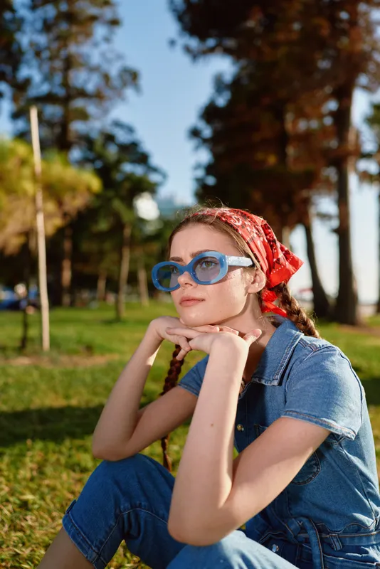 Lifestyle woman wearing modern boho-western denim street style with blue sunglasses and red bandana, sitting outdoors in soft film color light during sunny day.