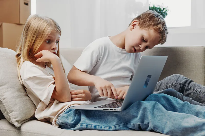 Two children sitting on a couch using a laptop together in a bright living room. Casual clothing, focused expressions, natural light creates a cozy home atmosphere for learning and play.