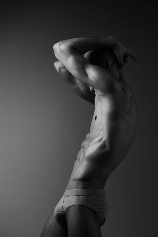 Black and white image of a man posing artistically, showcasing strong muscles and intense expression, emphasizing form and movement in a dramatic way.