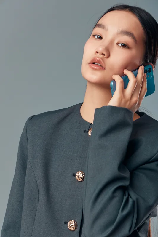Young woman posing with smartphone near ear wearing buttoned jacket, looking at camera with calm expression on neutral background, casual technology communication concept.