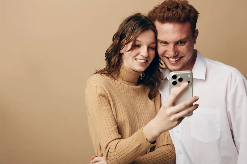 Smiling young couple takes selfie with smartphone on beige background. Casual clothing, happy mood, close relationship, technology use, modern lifestyle, digital communication concept.