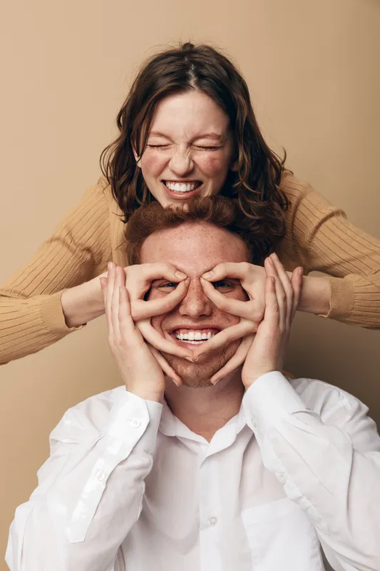 Happy young couple having fun, woman playfully covering man’s eyes with hands, both smiling and enjoying joyful moment together on beige background.