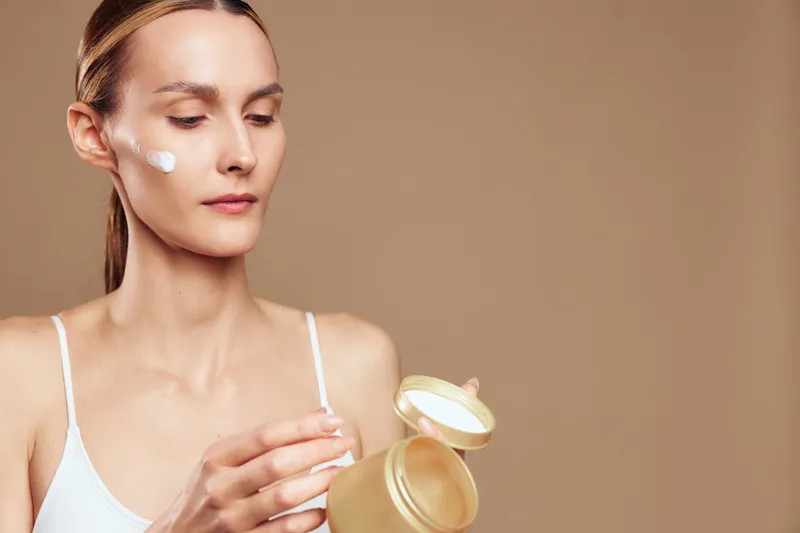 Young woman applying face cream with a calm expression, skincare routine, smooth skin, natural beauty, beige background, soft lighting, wellness concept