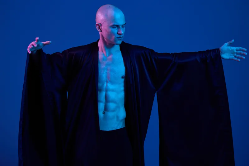 Strong man in black robe with a confident pose, showcasing powerful physique against a vibrant blue background. Unique concept reflects strength and artistry.